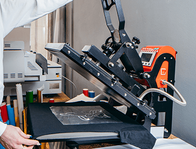  A heat press machine in operation, transferring a white design onto a black t-shirt in a professional printing workshop.