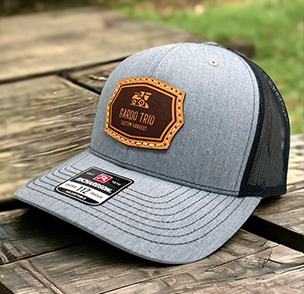 Grey and brown trucker hat with 'El Gordo Trio' patch, black mesh back, on a wooden surface.