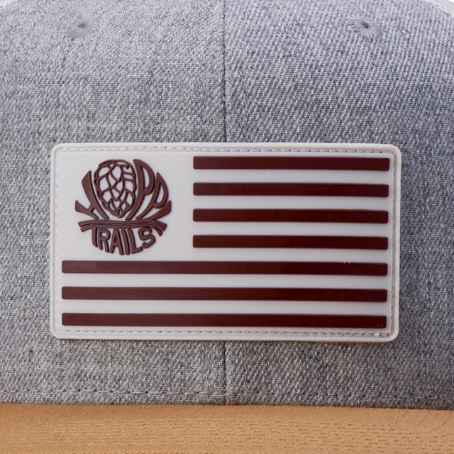 White rectangular rubber patch with "TRAILS" and pinecone emblem on a gray cap