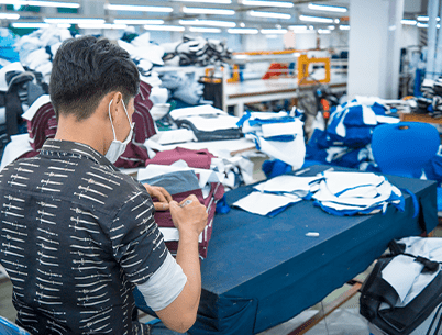 A masked employee inspecting fabric in a busy garment factory.