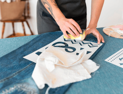 Person applying a stencil design on denim fabric for custom embroidery, with a sponge and paint, in a craft workspace.