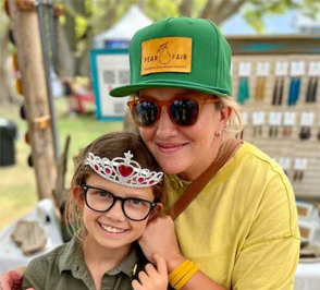 A woman and a girl with glasses pose together at a fair, both smiling. The woman wears a green cap with a PEAR FAIR patch