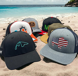 Assorted caps with various logos and designs, including an American flag, placed on a sandy beach.