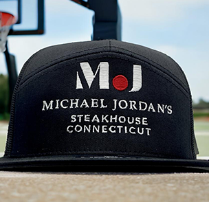 Black cap with 'MJ' logo and 'Michael Jordan's Steakhouse Connecticut' text, foreground focused.
