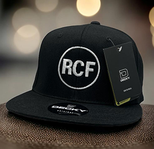 Black flat brim cap with bold 'RCF' letters, price tag attached, on a bokeh light background.