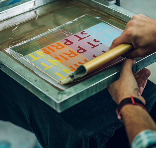Close-up of hands using a squeegee on a screen printing frame with colorful paint.