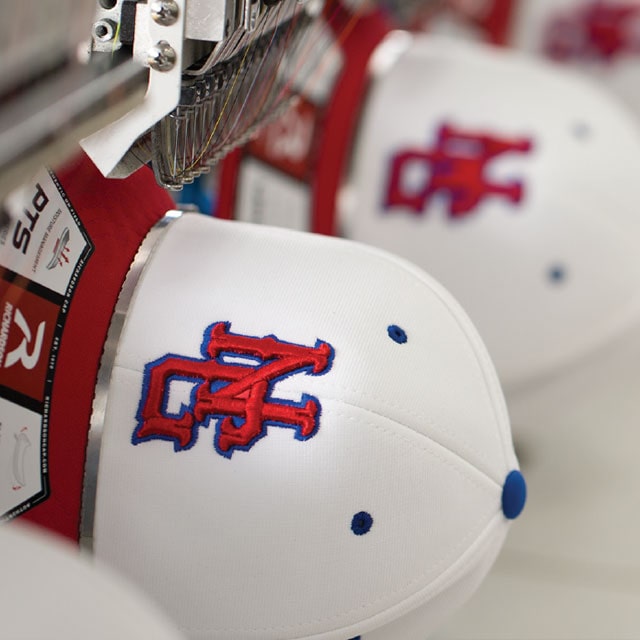 Embroidery machine stitching red and blue "OAC" team letters on a white cap