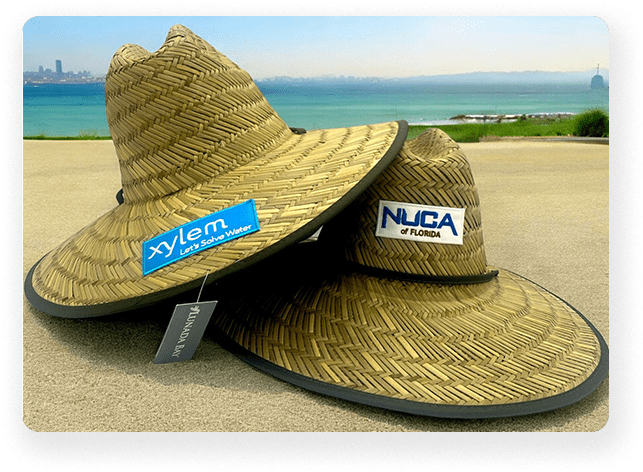 Two straw hats with custom patches, one labeled 'Xylem Let's Solve Water' in blue, and the other 'NUCA of Florida' in white, displayed outdoors by the water.